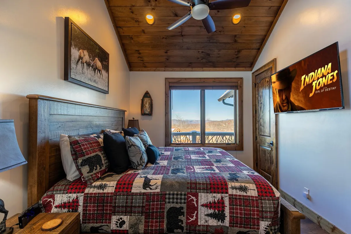 A cozy king bedroom with a sturdy wooden bed frame, a patterned quilt, a large window framing tranquil mountain and forest views, and a wall-mounted TV. Sunlight filters in, enhancing the serene, rustic atmosphere.