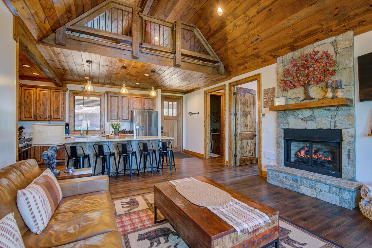 A spacious, open-concept living area featuring a cozy fireplace, a comfortable sofa, a modern kitchen island with bar seating, wood beams, and decorative wall art. The space is filled with natural light, and the layout allows for effortless flow between the living room, kitchen, and dining area.