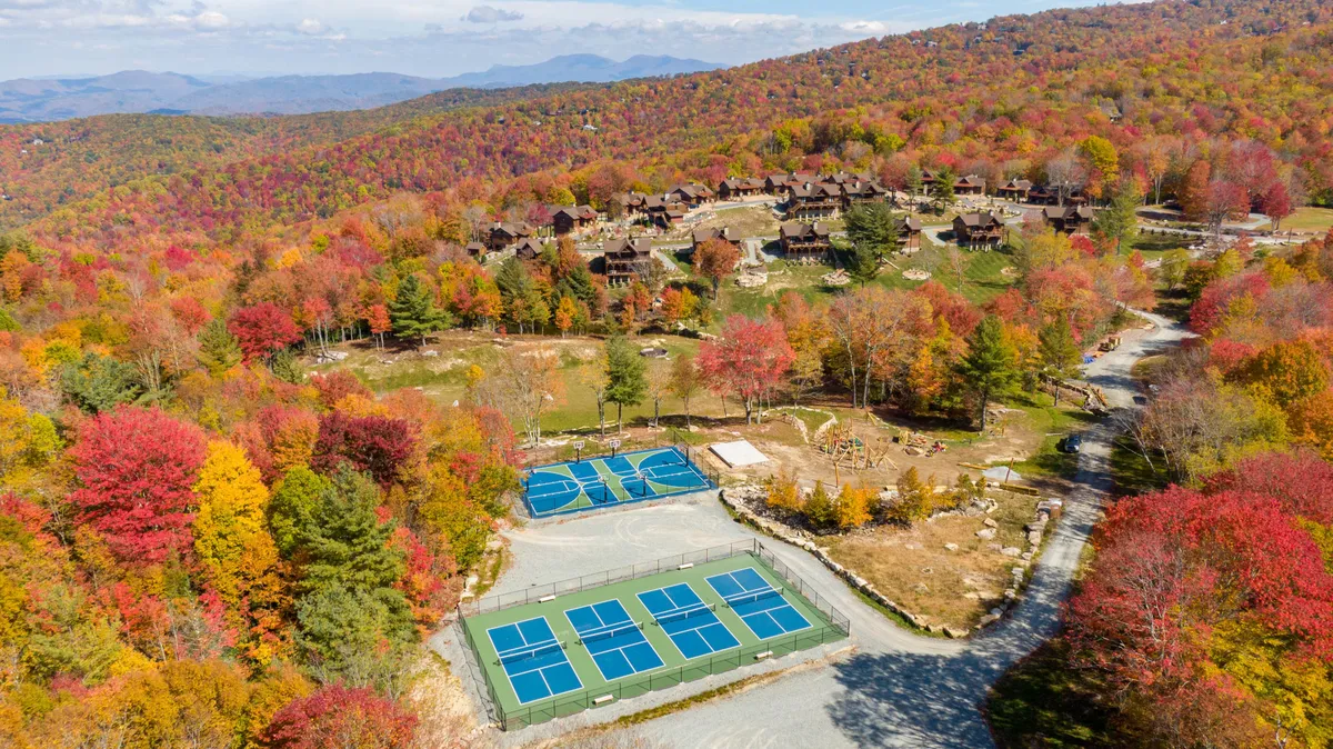 An energetic exterior view showing tennis courts surrounded by colorful autumn foliage, family-oriented lodging units, and a nearby playground. The scene feels lively, capturing recreational opportunities and the beauty of the resort setting.