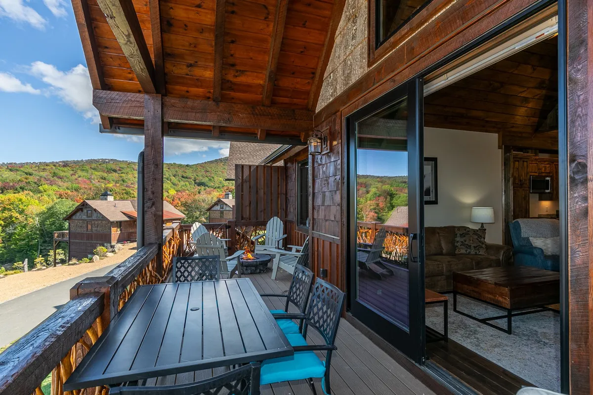A broad wooden deck is furnished with outdoor seating around a fire pit, looking out over sweeping mountain views. The expansive panorama is visible beyond the deck’s railing, suggesting a seamless connection between the interior living area and the outdoors. The inviting setting is ideal for socializing or stargazing by the fire.
