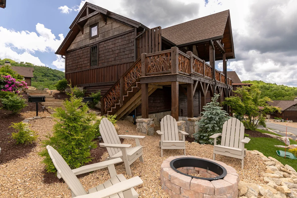 The image showcases a charming wooden cabin surrounded by a beautifully landscaped garden, with Adirondack chairs encircling a glowing fire pit. The scene captures a breathtaking sunset over the expansive mountain range, inviting guests to relax outdoors, enjoy nature, and gather with friends or family in a warm, inviting atmosphere.