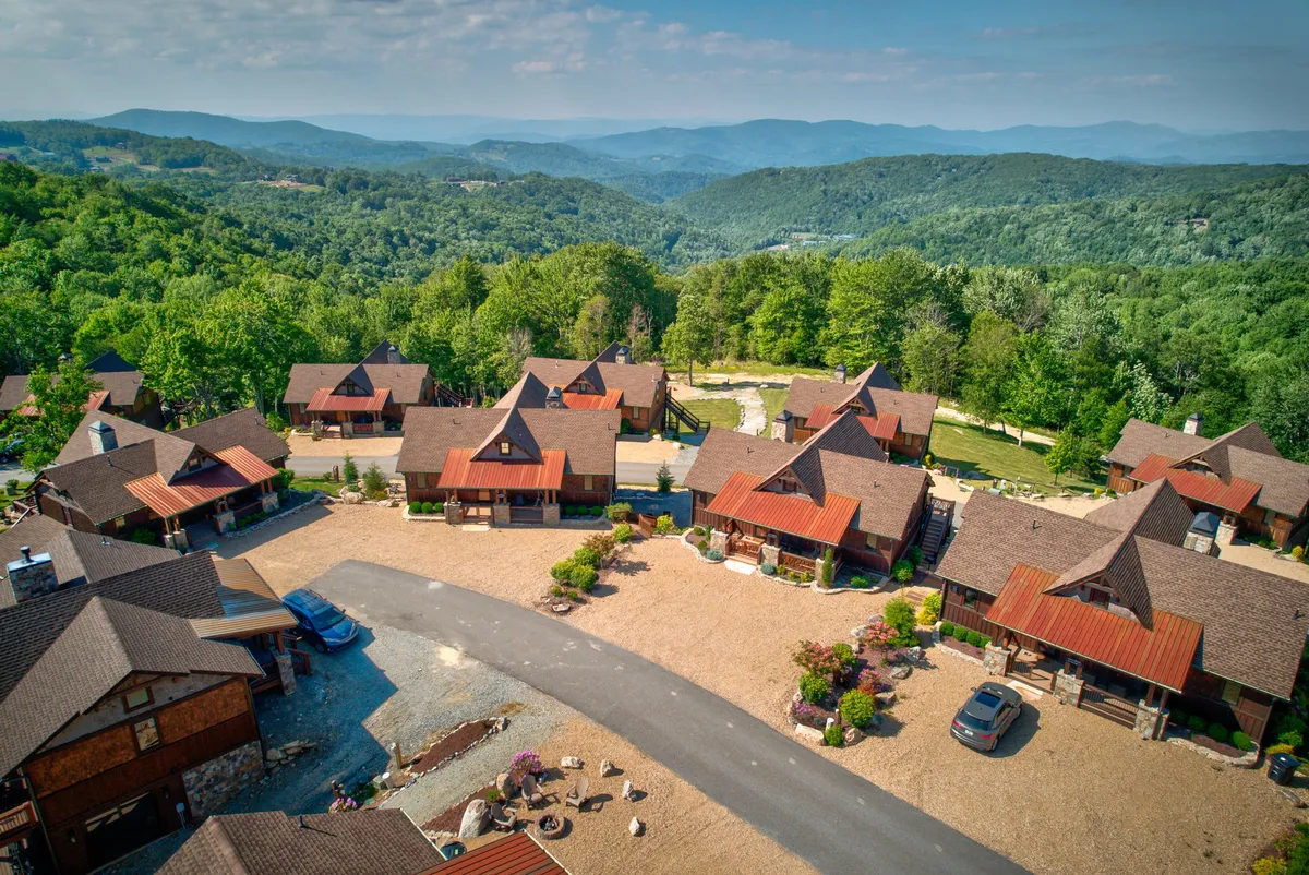 Multiple cozy cabins are set against a stunning mountain vista, with landscaped gardens and a driveway leading toward the wilderness. The image evokes a sense of tranquility while hinting at communal outdoor activities like barbecues and stargazing after an active day.