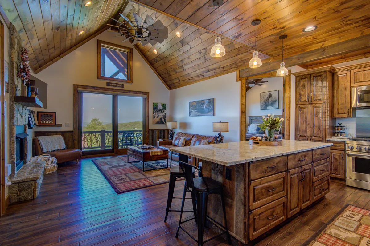 A warm, inviting living area with a large kitchen island at the center, a striking stone fireplace, sliding glass doors revealing expansive mountain views, and a comfortable seating area. The space is ideal for gatherings and offers abundant natural light.