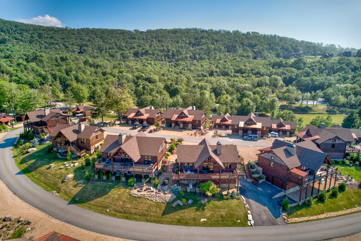 A series of log cabins lines a winding road, with landscaped gardens and tall trees creating a tranquil, inviting setting. The scene suggests easy access to nearby hiking and biking trails as well as space for outdoor gatherings.