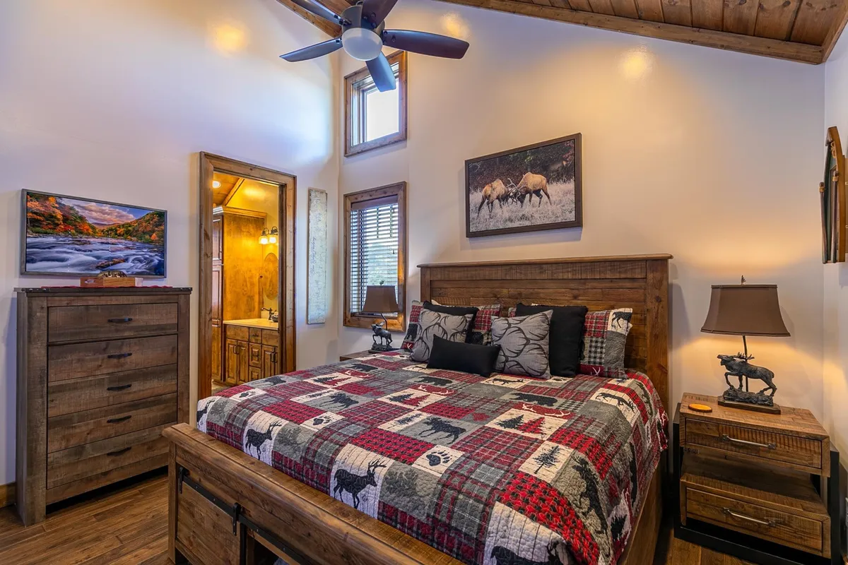 A warm, rustic bedroom featuring a bed with a patterned quilt, wooden dresser, wall artwork, and nightstands with soft lighting. The space radiates comfort and seclusion, ideal for restful nights.