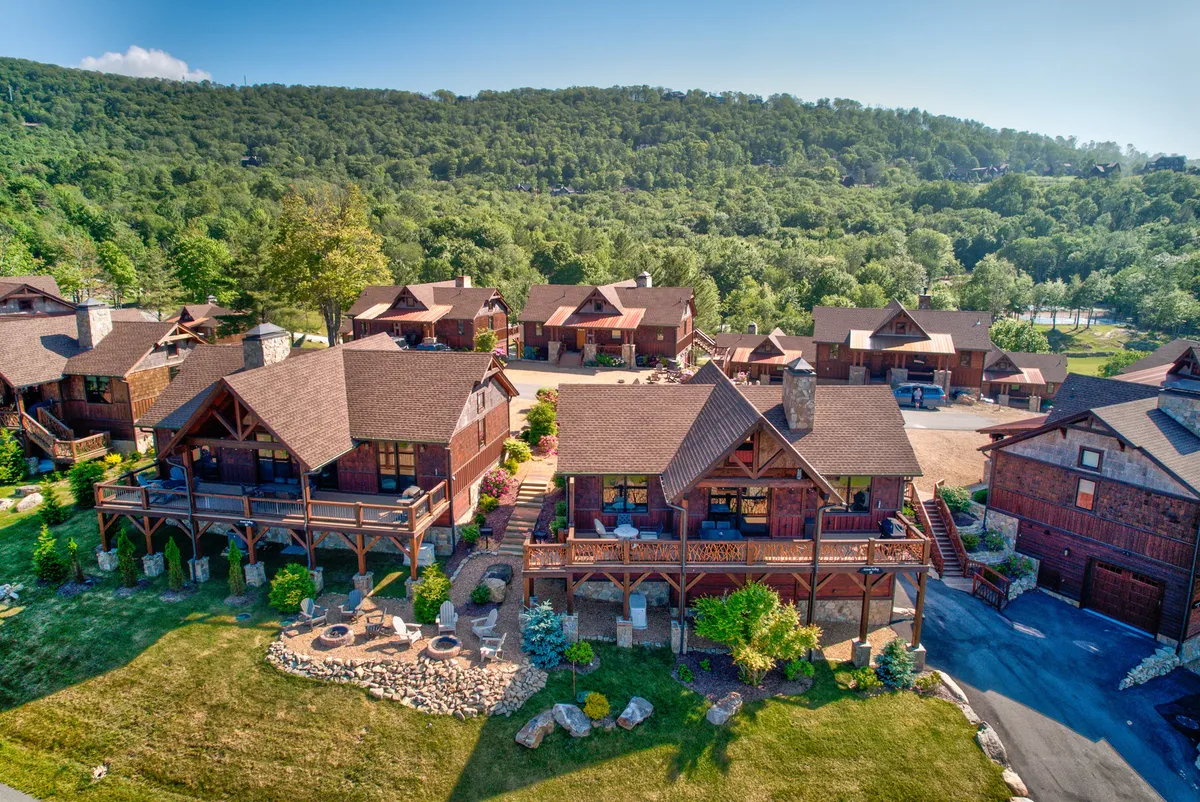 A charming exterior scene featuring log cabins, lush gardens, and stone pathways meandering through trees. The communal, open feel and visible porches evoke a friendly, outdoorsy atmosphere.